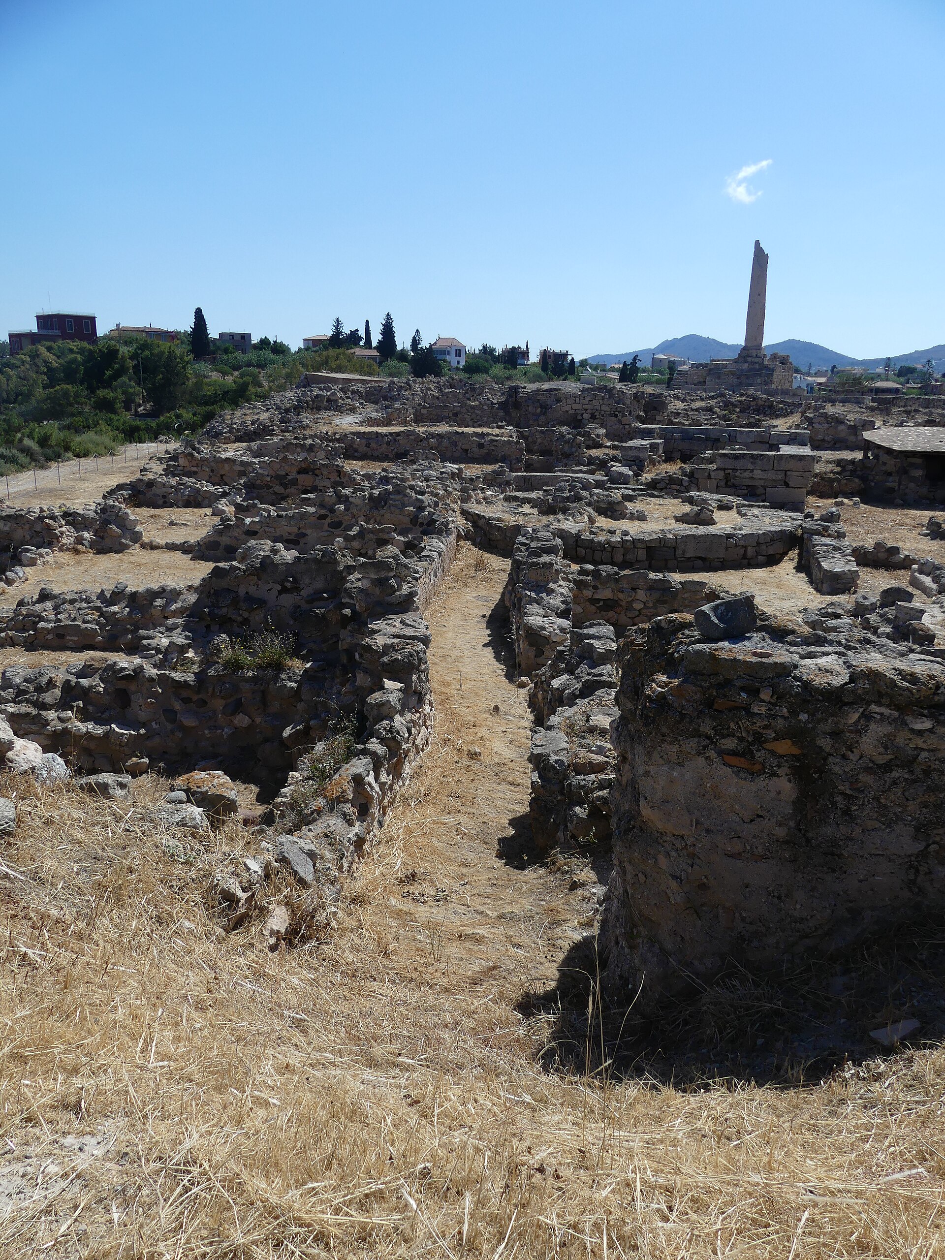 Low stone walls of Bronze Age buildings and a street at Kolona, with a single ancient column in the background